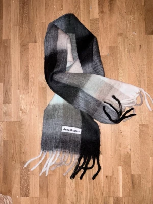 Rutig Acne Studios ullhalsduk - Säljer en fluffig och stor halsduk från Acne Studios i rutigt mönster. Färgerna går i svart, grått, beige och vitt. Halsduken har långa fransar i ändarna och en tydlig Acne Studios-label. Perfekt accessoar för att lyfta din outfit.