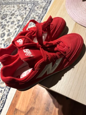 Röda New Balance RC42 sneakers - Säljer ett par röda New Balance RC42 sneakers med vit logga på sidan och gummisula. Skorna har snörning och är tillverkade i mocka och syntetmaterial. Snygg sportig look med rund tå och platt sula, perfekt för dig som gillar färgstarka sneakers.