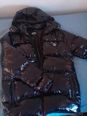 Svart pufferjacka från Trapstar - Säljer en glansig svart pufferjacka från Trapstar med stor huva och broderad logga på ryggen. Jackan har dragkedja framtill, fickor på sidorna och en extra ficka med knapp på bröstet. Perfekt för kalla dagar och riktigt snygg streetstil.