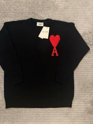 Svart stickad tröja från Ami Paris - Säljer en svart stickad tröja från Ami Paris med ikoniskt rött hjärta och A på bröstet. Tröjan har rund halsringning, långa ärmar och ribbade muddar. Perfekt för dig som vill ha en stilren men ändå statement look.