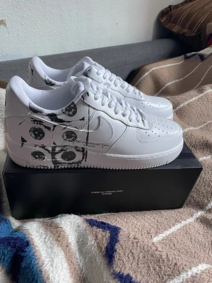 Nike Air Force 1 Supreme - Unika Nike Air Force 1 sneakers i samarbete med Comme des Garçons Shirt och Supreme. Skorna är vita i skinn med svart grafiskt ögonmönster på sidan. Klassisk låg siluett, perforerad tå och snörning. Perfekt för dig som vill sticka ut med en limiterad collab.