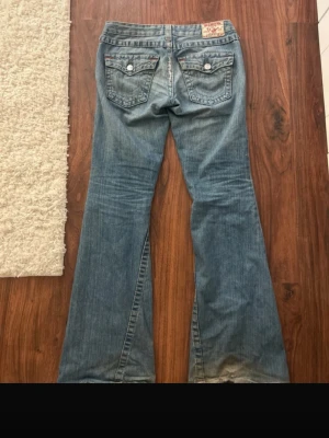 True Religion lowwaist bootcut jeans - Säljer ett par ljusblå bootcut jeans från True Religion med slitningar och hål på låret. Jeansen har dekorativa ficklock med knappar bak, klassiska kontrastsömmar och detaljerade sömmar på bakfickorna. Tillverkade i jeansmaterial med låg midja. Lite trasiga längst ner. Storlek w28