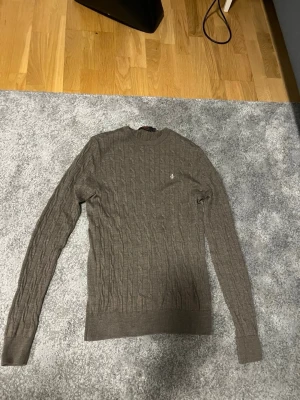 Brun kabelstickad tröja Morris S - Säljer en brun kabelstickad tröja från Morris i storlek S, tillverkad i 100% merinoull. Tröjan har långärmad design, rund hals och ett diskret Morris-emblem broderat på bröstet. Perfekt för dig som gillar klassisk stil med snygga detaljer.