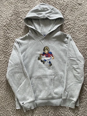 Polo ralph lauren hoodie - Säljer en klassisk grå hoodie från Polo Ralph Lauren med den ikoniska Polo Bear i amerikansk fotbolls-outfit på bröstet. En tidlös favorit som passar till det mesta! Är dock använd en del och därav priset (se sista bilderna). Hör av er vid föer frågor! 🤗