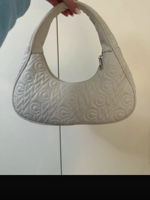Vit quiltad axelväska från CW - Säljer en snygg vit axelväska från CW med quiltat mönster där CW-loggan är insydd över hela väskan. Väskan har en mjuk, rundad form och stängs med dragkedja upptill. Perfekt storlek för det viktigaste och ger en clean look till din outfit.