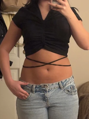 Croppad topp - Säljer en svart croppad topp från H&M Divided i storlek M. Toppen har korta ärmar, ribbad struktur och snygg snörning som går runt midjan och knyts i ryggen. V-ringad framtill med rynkade detaljer som ger en cool look.