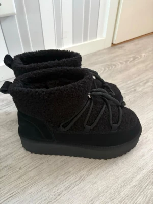 Svarta fluffiga boots från Duffy - Mysiga svarta boots från Duffy med fluffig utsida och tjock sula. Skorna har snörning framtill och är perfekta för kalla dagar. Materialet är en mix av syntet och mocka, vilket ger en mjuk och varm känsla. Rund tå och platt sula för extra komfort.