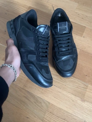 Valentino Garavani svarta sneakers - Säljer ett par svarta Valentino Garavani sneakers med coola camo-detaljer. Skorna har snörning, platt sula och blandning av mocka och skinn. Märkeslogga på plösen och nitar på hälen ger extra edge. Perfekta för dig som gillar exklusiva och stilrena sneakers.