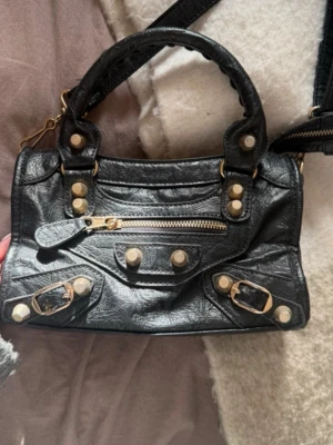 Svart handväska med gulddetaljer - Snygg svart handväska i skinnimitation med guldfärgade nitar och spännen. Väskan har både handtag och axelrem samt en dragkedja framtill. Perfekt storlek för det viktigaste och ger en cool vibe till din outfit.