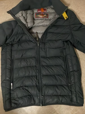 Svart pufferjacka från Parajumpers S - Säljer en svart pufferjacka från Parajumpers i storlek S. Jackan är superlätt, har quiltad design, dragkedja framtill och två sidofickor med dragkedja. Insidan är grå och jackan har en gul detalj vid kragen. Perfekt för kalla dagar när du vill hålla stilen.
