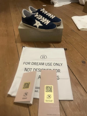 Blå sneakers från Golden Goose - Säljer ett par snygga blå sneakers från Golden Goose med vit stjärndetalj på sidan och guldfärgad häl. Skorna har vita snören och en klassisk låg siluett. Ovandelen är i mocka och sulan är platt. Perfekta för dig som gillar streetstyle och vill ha något unikt.