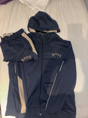 Hugo boss set - Säljer ett par marinblå mjukisbyxor från Hugo Boss med beigea detaljer längs benen och logga på ena låret. Byxorna har resår i midjan och är tillverkade i mjukt material som känns skönt mot huden. Perfekta för chill dagar eller när du vill ha en sportig look.