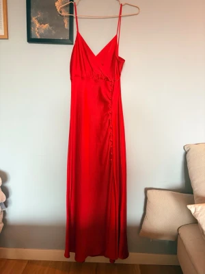Röd långklänning från Zara storlek M  - Säljer en röd långklänning med smala axelband och omlott-vringning. Klänningen har dekorativa knappar längs sidan och är gjord i ett glansigt, mjukt material som ger en lyxig känsla. Passformen är lite åt det mindre hållet mer som en S jag som referens är 157 och storlek XS/S. Har lite trådning se bilder men inget som påverkar funktionen eller håll eller liknande. Accepterar prisförslag 