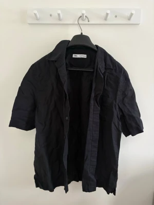 Herr Svart linneskjorta från Zara  - Svart skjorta från Zara i storlek S, tillverkad i linne. Skjortan har klassisk krage, korta ärmar och knäppning framtill med svarta knappar. Perfekt för dig som gillar enkel och stilren design.