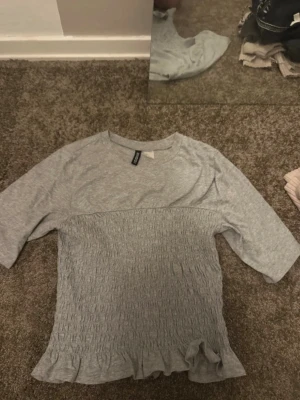 Grå ribbad topp från H&M Divided - Säljer en ljusgrå topp från H&M Divided med ribbad, smockad midja och volangkant nedtill. Toppen har rund halsringning och trekvartsärm. Perfekt till jeans eller kjol för en chill look.