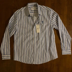 Randig skjorta  - Snygg skjorta  i vitt och ljusblått med vertikala ränder. Klassisk krage, långa ärmar och knäppning framtill. Broderad A-logga på bröstet. Tillverkad i mjuk bomull för en clean och fräsch look. Den har ingen etikett på sig längre och storleken är som en S