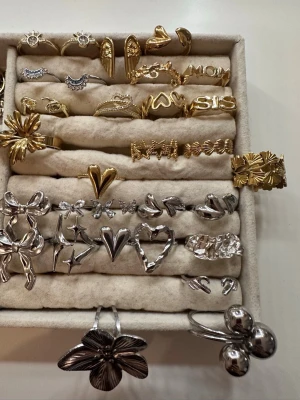 Ringar  - Bilden visar ett urval av ringar i guld och silver med olika former som hjärtan, blommor, rosetter och text. Ingen väst finns på bilden.