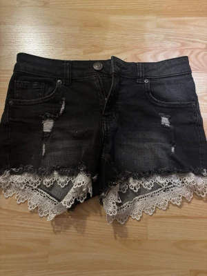 Svarta jeansshorts med spets från Amisu - Svarta jeansshorts från Amisu i storlek 34. De har slitna detaljer, fransig kant och vit spets som ger en snygg kontrast. Perfekta på sommaren 