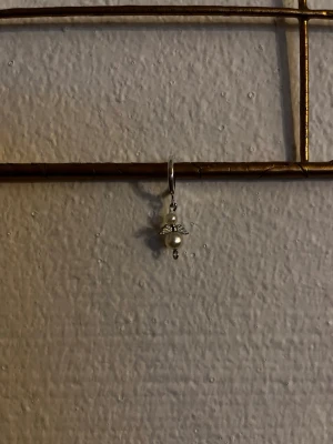 Pärlörhänge med ängelberlock - Unikt örhänge i silverfärg med en söt ängelberlock. Berlocken består av två vita pärlor och små detaljerade vingar i metall. Perfekt accessoar för dig som gillar personliga smycken med en touch av elegans.