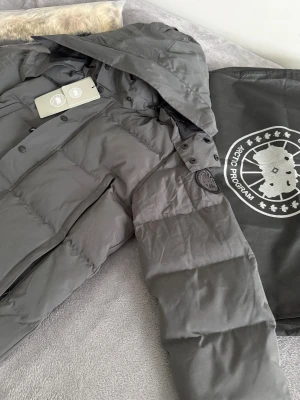 Canada Goose jacka  - Säljer en mörkgrå dunjacka från Canada Goose med huva och klassisk svart logotyp på ärmen. Jackan har raka linjer, stora fickor med dragkedja och knappar framtill. Medföljer en lös pälskant till huvan. Perfekt för kalla dagar.