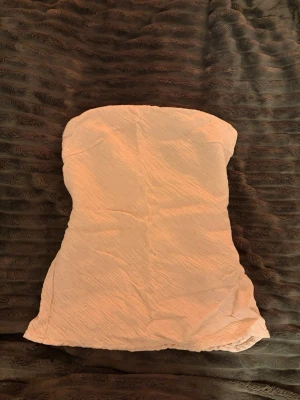 Beige tubtopp från Lindex XS - Säljer en beige tubtopp från Lindex i storlek XS. Toppen är axelbandslös och har en lätt skrynklig struktur i tyget som ger en snygg effekt. Perfekt till jeans eller kjol för en enkel och stilren look.