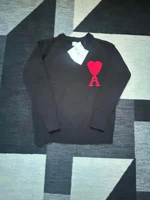 Ami Paris stickad Tröja-helt ny  - Säljer en svart stickad tröja från Ami Alexandre Mattiussi med röd hjärtlogga och bokstaven A på bröstet. Tröjan har ribbade muddar, rund hals och lång ärm. Perfekt för dig som vill ha en clean men ändå statement look. Kommer med original påsen. Kan diskutera pris.