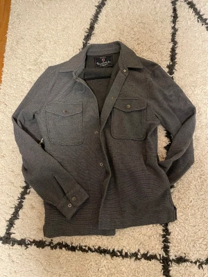 Overshirt från Bondelid  - Snygg grå overshirt från Bondelid med diskret mönster och två bröstfickor med knappar. Klassisk krage, långärmad och med slitsar i sidorna. Perfekt att ha som lager över t-shirt. Modern och stilren look. Storlek S men sitter som S-M. Priset är inte hugget i sten!