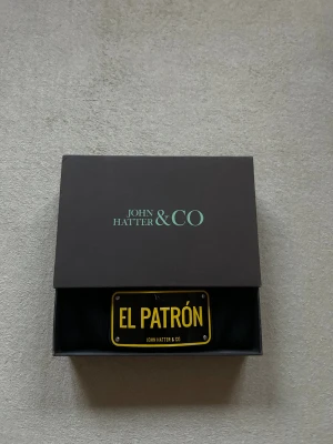 Svart mössa John Hatter & Co El Patrón - Svart mössa från John Hatter & Co med uppvikt kant och en cool metallskylt framtill med texten 'EL PATRÓN' i gult. Mössan har en klassisk form och är tillverkad i mjukt stickat material. Perfekt för dig som vill sticka ut med en unik detalj.