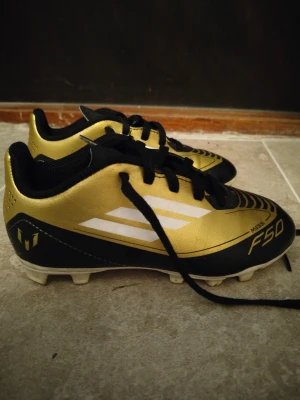Adidas Messi F50 fotbollsskor guld/svart - Säljer ett par Adidas Messi F50 fotbollsskor i guld och svart med vita detaljer. Skorna har snörning, platt sula med fasta dobbar och är tillverkade i syntetmaterial. Ikonisk Messi-logga på hälen och snygg design för dig som vill sticka ut på planen.