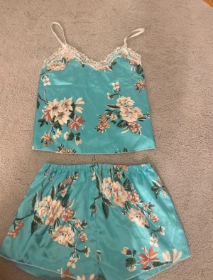 Pyjamas set - Säljer en turkos linnetopp med blommigt mönster och tunna axelband. Toppen har dekorativ vit spets längs halsringningen och är tillverkad i ett glansigt, mjukt material som känns lyxigt mot huden. Perfekt för dig som gillar färg och romantiska detaljer.