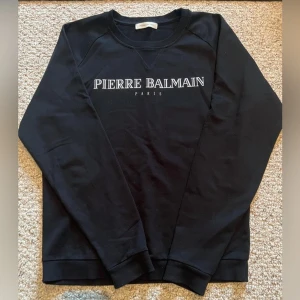 Svart Pierre Balmain sweatshirt storlek S - Svart sweatshirt från Pierre Balmain med vit logotyp på bröstet. Tröjan har rund hals, ribbade muddar och är tillverkad i 100% bomull. Långärmad och klassisk passform, perfekt för dig som gillar stilrena märkesplagg.