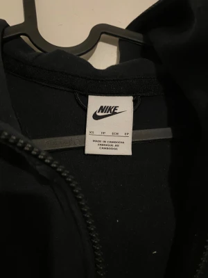 Nike tech fleece - Svart hoodiejacka från Nike med hel dragkedja och huva. Jackan har ett stilrent V-format band över bröstet och en ficka med dragkedja på ärmen. Nike-logga i grått på bröstet. Perfekt för chill eller streetwear.