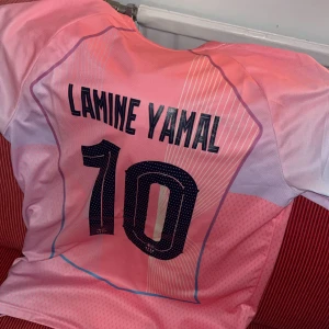 Barcelona Lamine Yamal #10 rosa tröja - Säljer en riktigt snygg FC Barcelona fotbollströja från Nike med Lamine Yamal och nummer 10 på ryggen. Tröjan är rosa med ljusblå detaljer, klubbmärke, Champions League-märke på ärmen och Spotify-logga på bröstet. Perfekt för dig som älskar Barca och vill sticka ut.