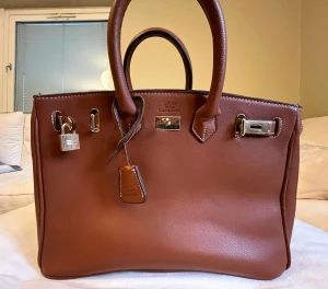 Birkin brun handväska i skinn - Exklusiv brun Birkin handväska (storlek 35)  i lyxigt skinn med guldfärgade metalldetaljer, lås och nyckel. Klassisk fyrkantig form med dubbla handtag och synliga vita sömmar. Perfekt för dig som vill ha en ikonisk och tidlös accessoar.