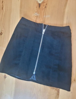 Svart kortkjol med dragkedja - Snygg svart kortkjol med en cool silverfärgad dragkedja framtill. Kjolen har en rak passform och är perfekt för dig som gillar en stilren och edgy look. Passar till många olika outfits och är lätt att matcha.