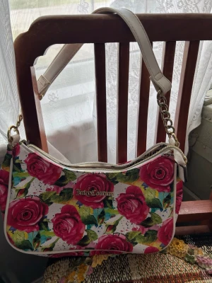 Blommig axelväska från Juicy Couture - Säljer en helt ny vit axelväska från Juicy Couture med färgglatt ros-mönster i rött, grönt och blått. Väskan har guldfärgade detaljer, kedja på axelremmen och dragkedja. Köpte den för 102$ (c 960kr) i USA. Säljer för 700kr. 100% äkta!! Aldrig använd.