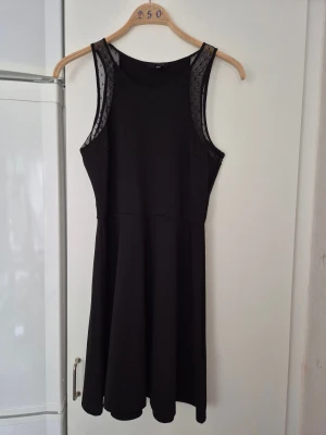 Klänning - Snygg svart klänning från H&M i storlek M. Modellen är ärmlös med mesh-detaljer vid axlarna och har en rundad halsringning. Kjolen är lätt utsvängd och klänningen har en enkel, stilren look som känns modern och fräsch.