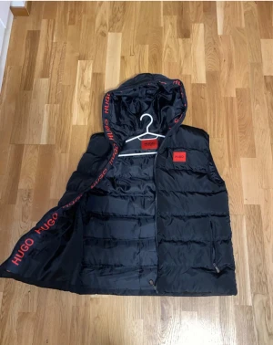 Hugo, väst - Svart dunväst från Hugo med röd logga på bröstet och röd text på insidan av huvan. Västen har endast använts en gång så prislapp finns inte kvar men västen är i nyskick! Västen är köpt utomlands för 2600:- Storlek M/L. Priset kan alltid diskuteras 😊