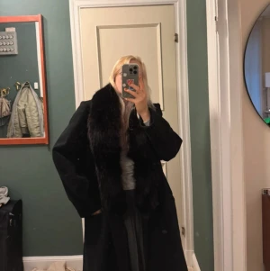 Svart lång kappa med pälskrage - Säljer en svart lång kappa med bred, fluffig päls runt kragen. Klassisk rak modell med långa ärmar och stilren design. Perfekt för dig som vill ha en elegant och tidlös look under kallare månader.