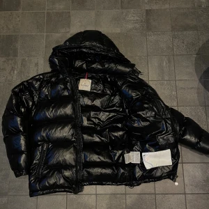Svart Moncler pufferjacka med huva - Säljer en svart pufferjacka från Moncler med glansig finish och huva. Jackan har dragkedja framtill, två fickor med dragkedja och klassisk Moncler-logga på insidan. Fylld med äkta dun för extra värme, perfekt för kalla dagar.