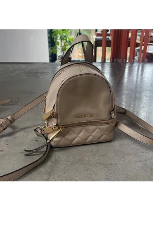 Beige quiltad ryggsäck Michael Kors - Snygg liten beige ryggsäck från Michael Kors i skinn med quiltad nederdel och guldfärgade detaljer. Väskan har flera fack med dragkedja och justerbara remmar. Perfekt storlek för det viktigaste och stilren design med logga framtill.