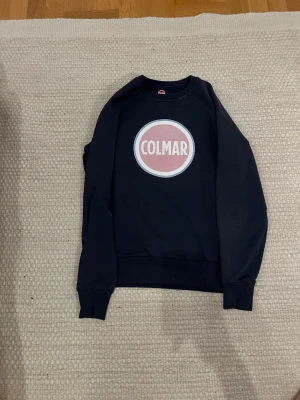Mörkblå Colmar sweatshirt med logga - Snygg mörkblå sweatshirt från Colmar i storlek M. Tröjan har ett stort, rundat Colmar-tryck i vitt och rött på bröstet. Klassisk passform med ribbade muddar vid ärmar och nederkant. Tillverkad i mjuk bomull som är skön mot huden.
