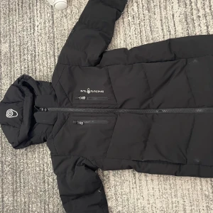 Svart dunjacka från Sail Racing - Svart dunjacka från Sail Racing med huva och två bröstfickor med dragkedja. Jackan har broderad logga på bröstet och quiltad design. Materialet är polyester och dun, perfekt för kalla dagar. Snygg och stilren look med lång ärm och normal passform.