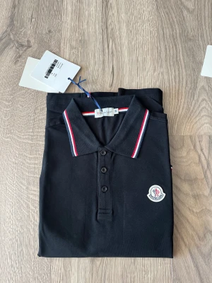 Svart Moncler pikétröja med röd/vit detalj - Svart pikétröja från Moncler med klassisk krage och knappslå. Tröjan har Moncler-logga broderad på bröstet och snygga rödvita ränder på krage och ärmslut. Tillverkad i bomull och har korta ärmar, perfekt för en clean och sportig look.