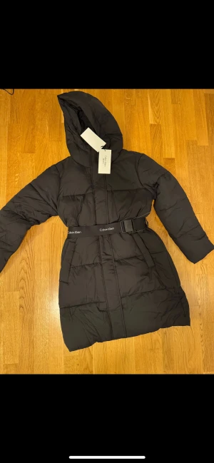 Svart pufferjacka från Calvin Klein - Svart lång pufferjacka från Calvin Klein  med huva och justerbart bälte med logga i midjan. Jackan har dragkedja framtill och är quiltad för extra värme. Perfekt för kalla dagar och har en clean, modern look. Prislappen är kvar  med original pris som va 1450kr