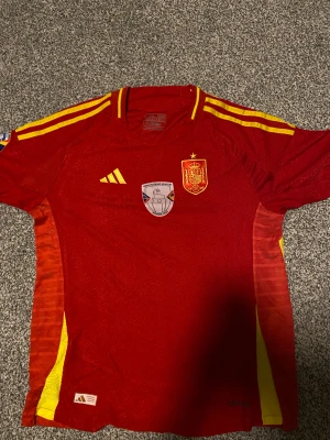 Spaniens Euro 2024 tröja - Säljer en officiell röd Spanien fotbollströja från Adidas. Tröjan har gula detaljer, broderat landslagsmärke och Nations League patch på bröstet och ärmen. Tillverkad i ventilerande polyester med korta ärmar och slim fit. Storleken är tajt så passar för XL eller L
