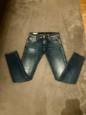 Replay Anbass slim fit jeans blå - Snygga Replay Anbass jeans i slim fit med klassisk femficksdesign och coola slitningar. Färgen är mörkblå med ljusare tvättade partier. Bakfickorna har kontrastsömmar och jeansen är tillverkade i återvunnet denimmaterial.