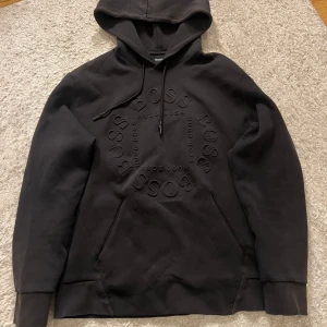 Svart Hugo Boss Hoodie - Svart hoodie från Hugo Boss med stor logga i cirkel på bröstet. Tröjan har luva med snören. Färgen har tappats lite men inget man märker. Pris kan sänkas vid snabb affär och hör av er vid intresse eller liknande!😁