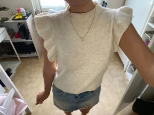 Vit stickad topp med volangärmar - Säljer en superfin vit stickad topp med volangdetaljer på axlarna. Toppen är ärmlös med rund hals och har en mjuk, lite fluffig känsla. Perfekt att styla med jeans eller kjol för en fräsch look. Passformen är normal och toppen är croppad.