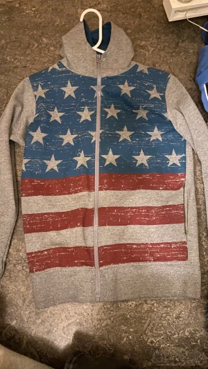 Grå hoodie med amerikanskt flaggmönster - Snygg grå hoodie från KappAhl i storlek 170, med dragkedja och huva. Framsidan har ett coolt amerikanskt flaggmönster i blått, rött och vitt med stjärnor och ränder. Materialet är mjukt och känns som bomull, perfekt för chill dagar och en cool outfit 